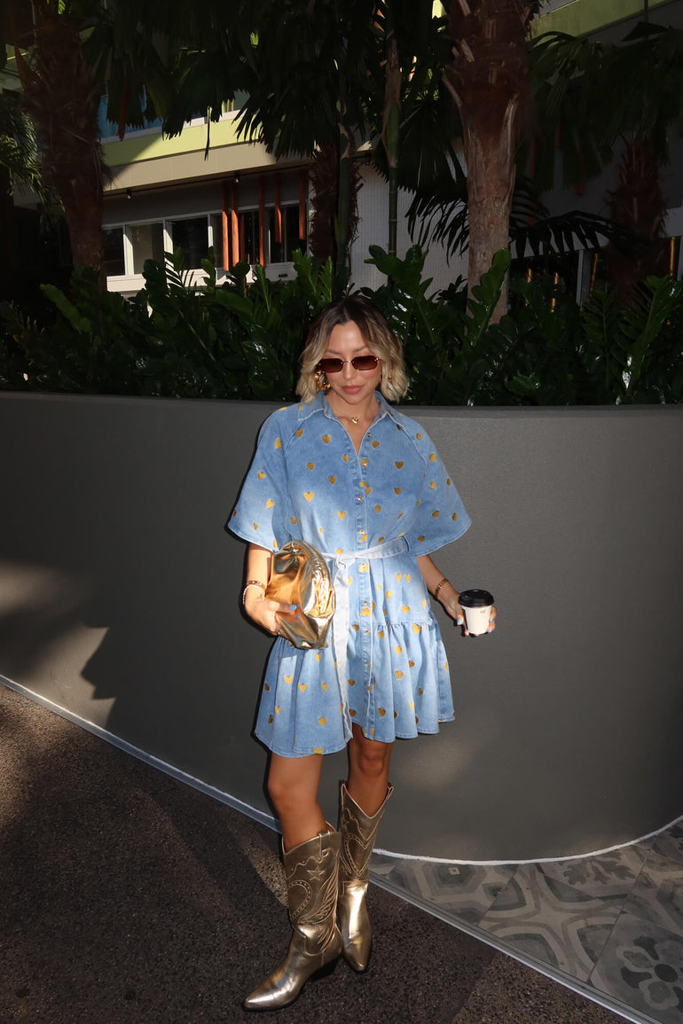 Woman in a blue dress with gold hearts and cowboy boots holding a clutch and cup outdoors