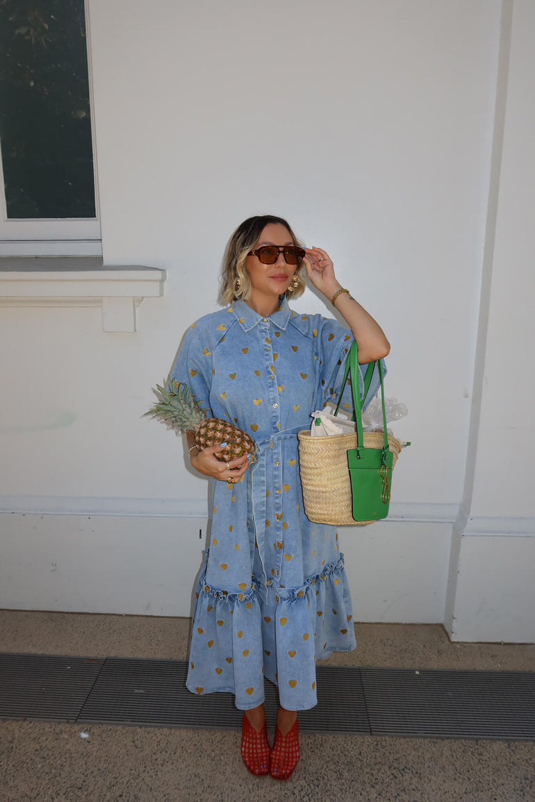 Woman in a blue dress holding a pineapple and a green bag against a white wall.
