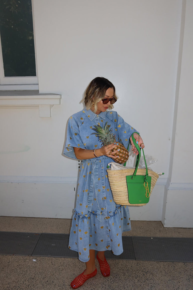 Woman in a blue dress holding a pineapple and a green bag against a white wall.