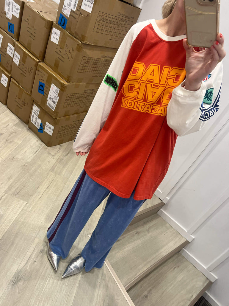 Person wearing a red and white sweatshirt with text, blue Terry towelling pants, and silver shoes in a room with cardboard boxes.