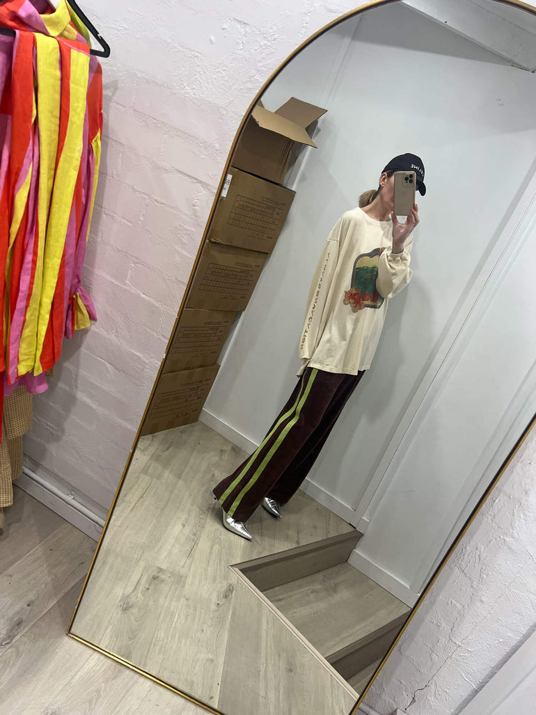 Person taking a mirror selfie wearing a beige sweater and striped Terry towelling pants in a room with colorful clothes on a rack.