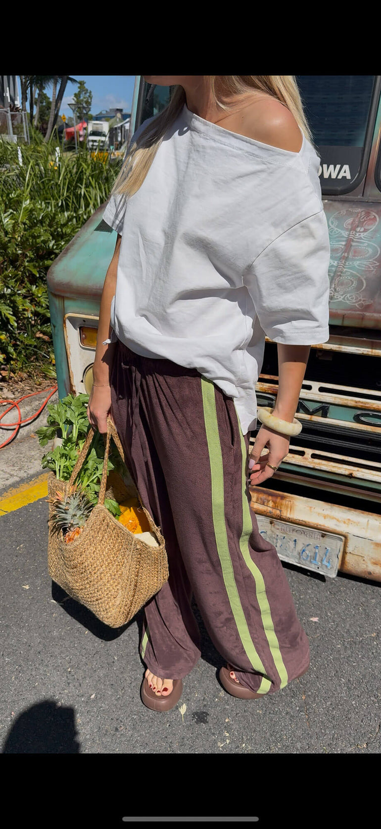 Person holding a woven bag with groceries, wearing a white top and brown terry towelling pants with green stripes, standing next to a vehicle.