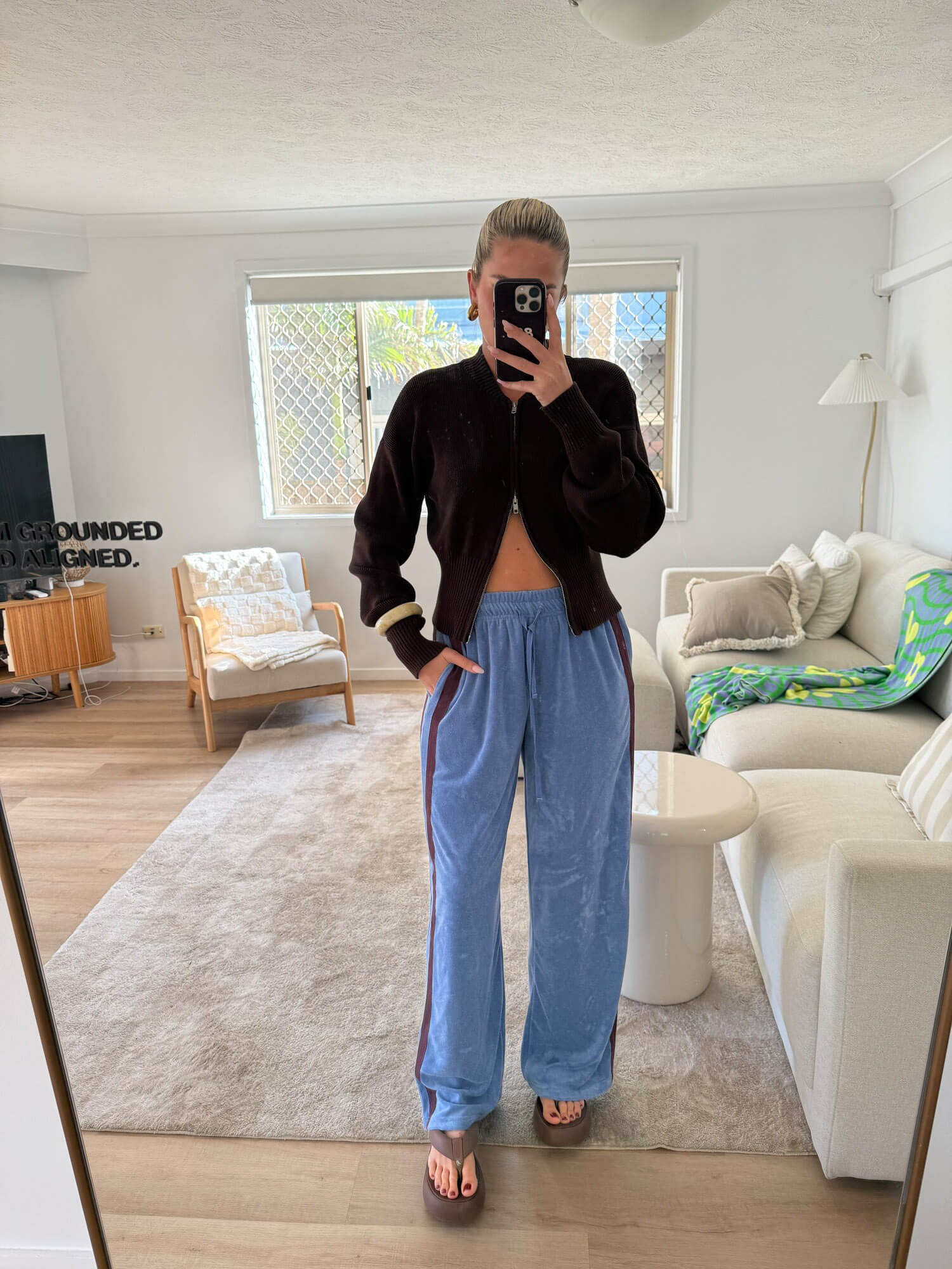 Person taking a mirror selfie in a living room wearing a black jacket and blue terry towelling pants.