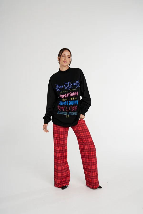 Person wearing a black Summi Summi sweater with colourful text and red plaid pants on a white background