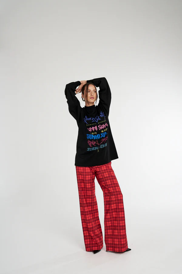 Person wearing a black Summi Summi sweater with colourful text and red plaid pants on a white background