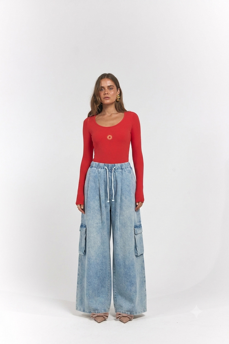 A woman stands against a light gray studio background wearing a bright red long-sleeve scoop-neck top and light-wash wide-leg denim cargo pants.