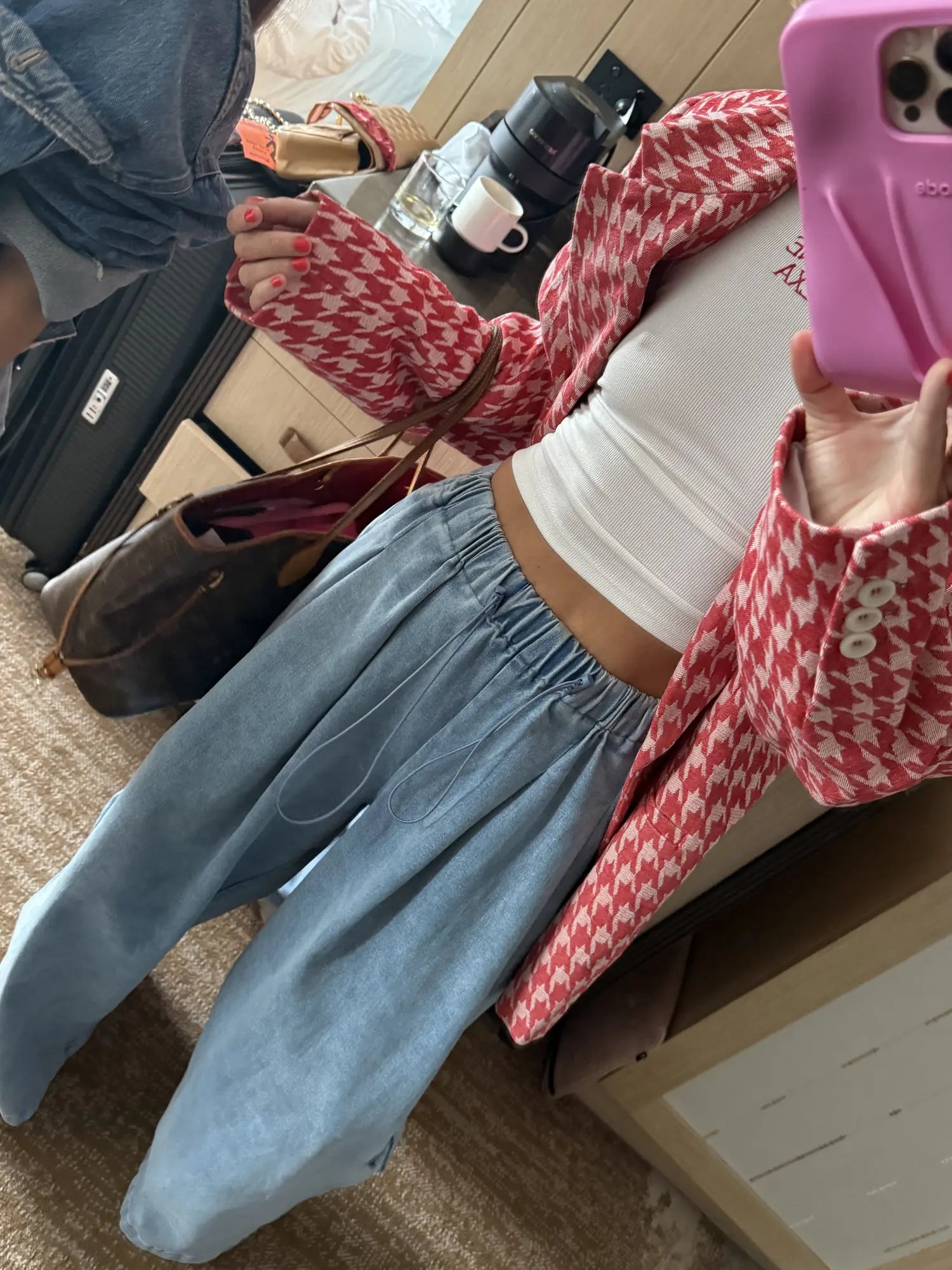 A person taking a mirror selfie in a bedroom setting, wearing a red and white houndstooth patterned cardigan over a white crop top and light blue wide-leg jeans.
