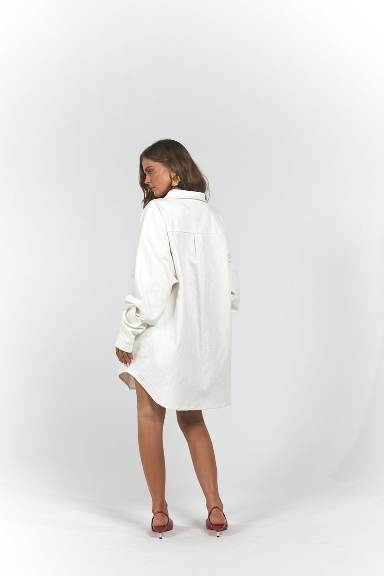 A woman wearing an oversized white shirt dress, shown from the back in a studio setting.