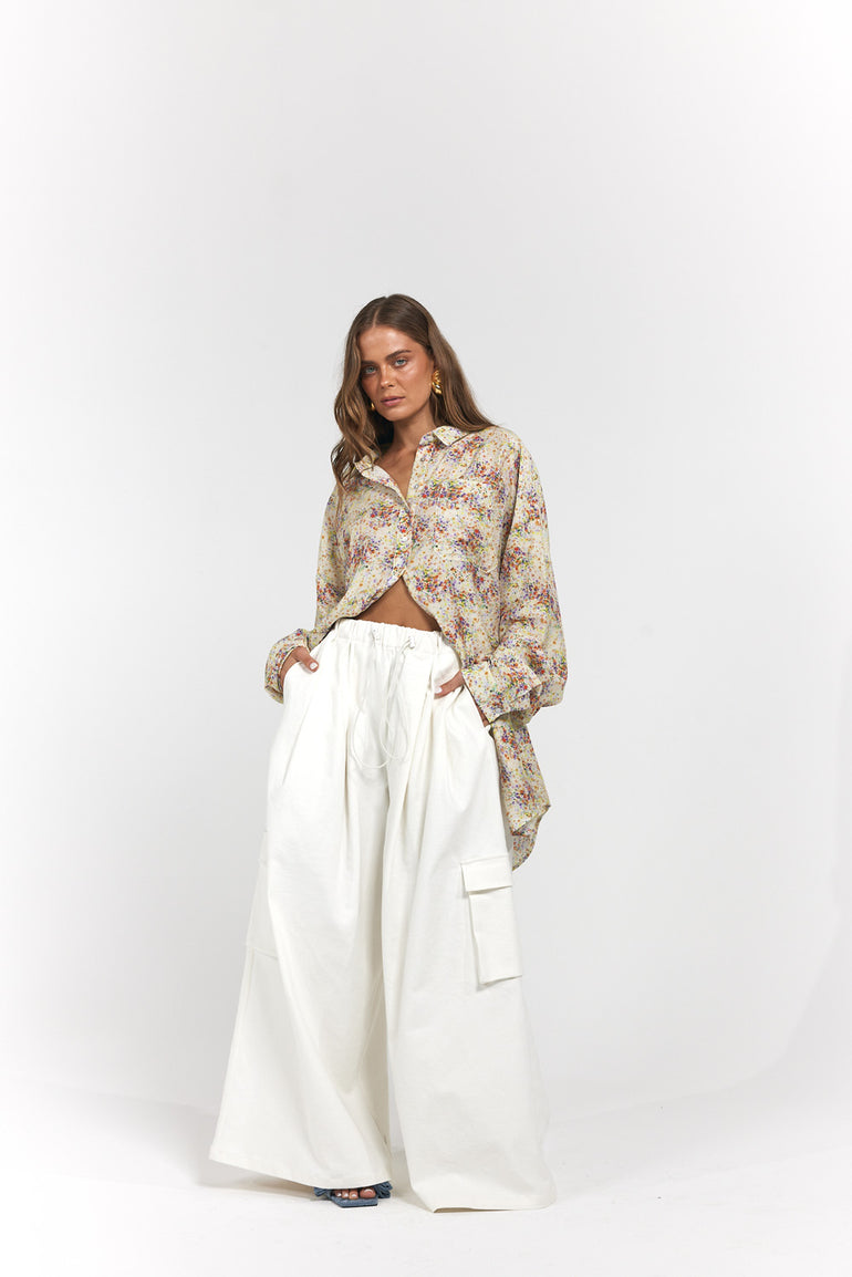 A woman modeling a long-sleeve button-up shirt with a beige, brown, and muted red floral pattern, paired with wide-leg white cargo pants.