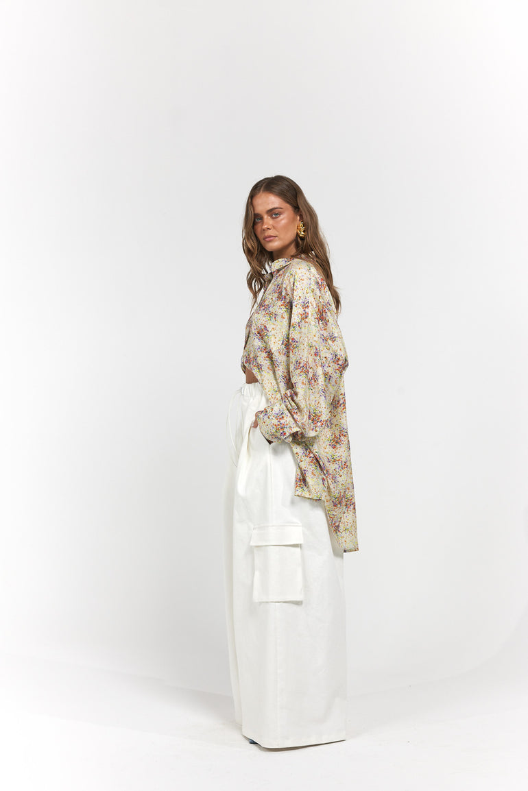 A model is shown from the side in a studio setting, wearing a cream-colored floral print shirt and a long white skirt with a large front pocket.