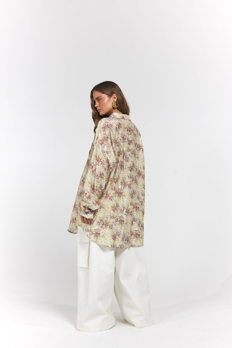 A woman wearing a loose, long-sleeve floral print shirt in beige with muted pink and green tones, paired with wide-leg white pants.