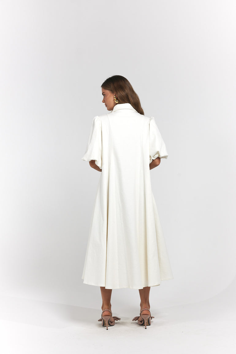 A woman viewed from the back wearing a long white midi dress with short puff sleeves and a high collar, paired with brown heeled sandals.