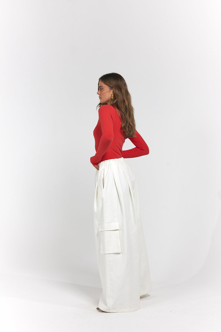 A woman standing in profile wearing a bright red long-sleeve top and high-waisted white wide-leg trousers with side cargo pockets.