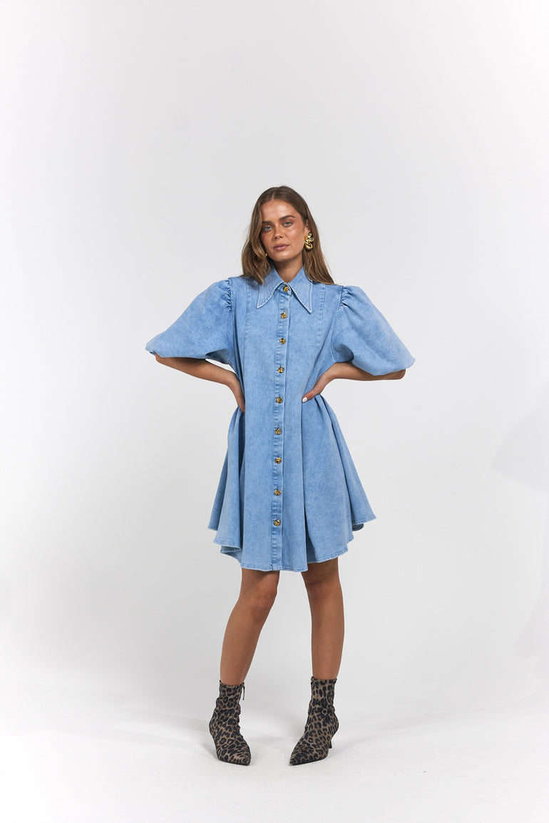 A woman modelling a light blue denim shirt dress with puff sleeves and a button-down front, paired with dark patterned ankle boots.